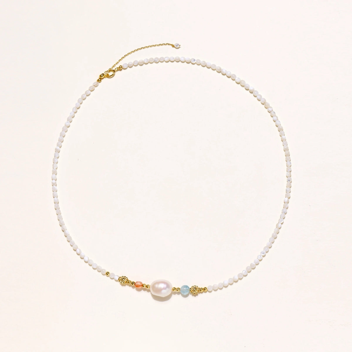 Delicate white beaded necklace with baroque pearl center, gold accents, and coral and aqua gemstones on cream background.